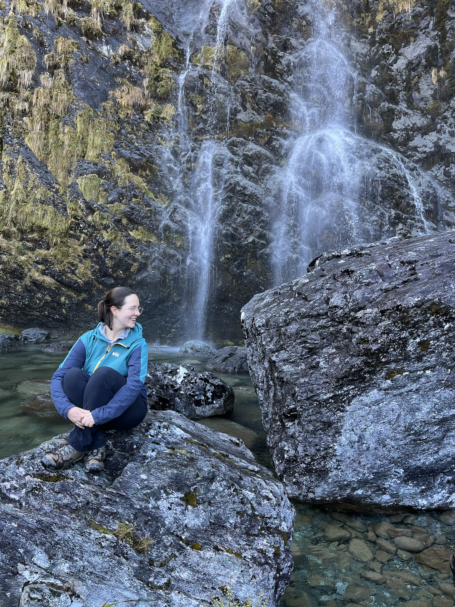 Image of a woman sitting on a rock. She is surrounded by water and there is a waterfall in the background. She looks happy.